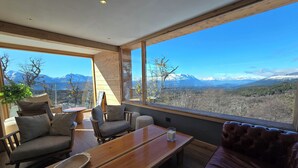 Una habitación moderna con una mesa de madera, sillas y un sofá. Amplios ventanales ofrecen vistas a montañas y árboles.