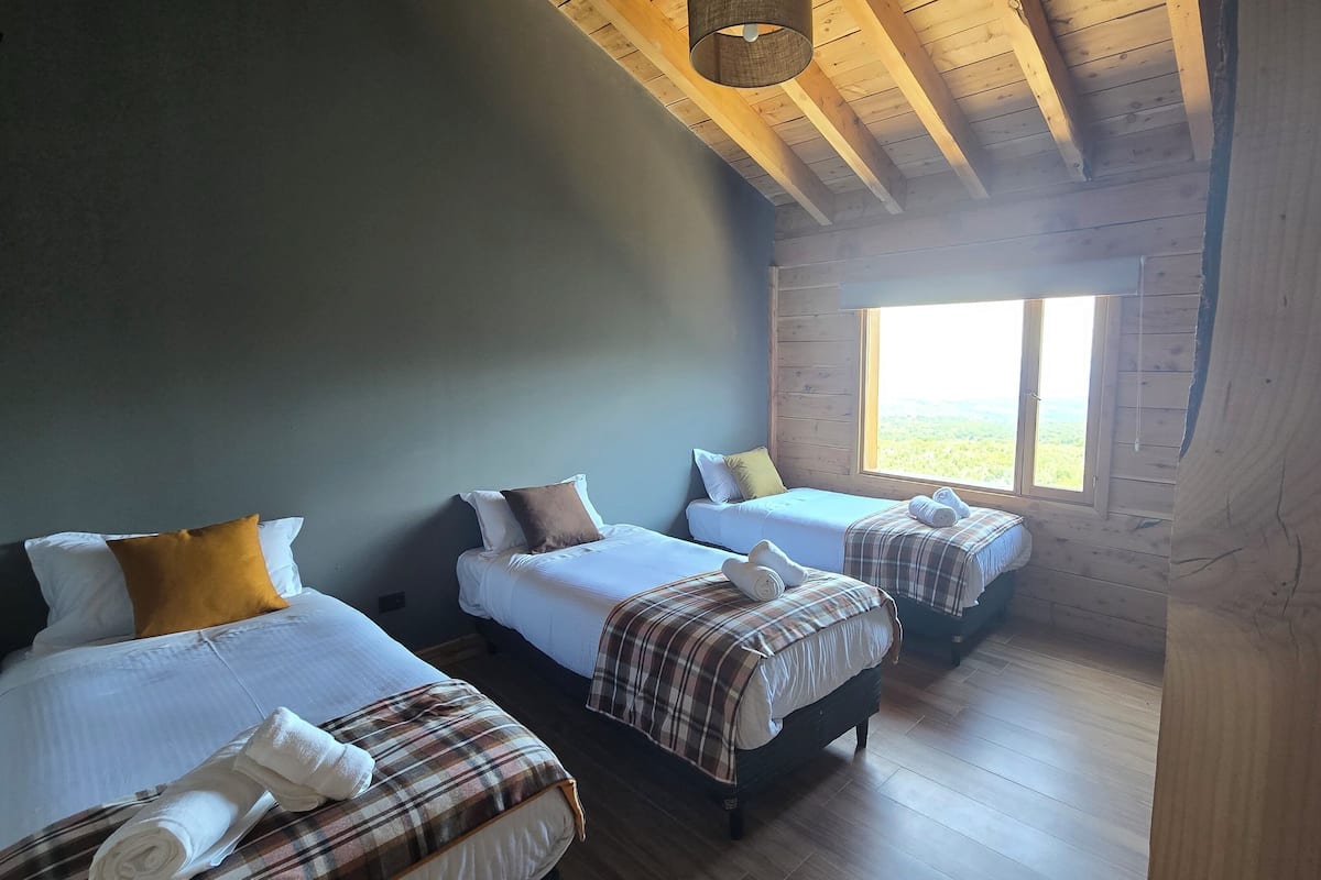 A room with two beds, wooden flooring, a large window, and a wooden ceiling.