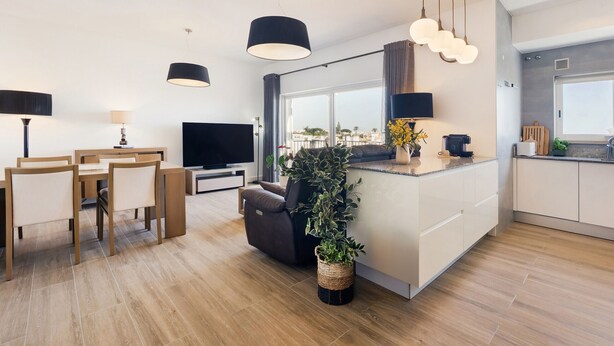 A modern living space with a dining area, sofa, TV, kitchen, and large windows. - Apt. Deluxe OCV - Pool & Albufeira View (Albufeira)