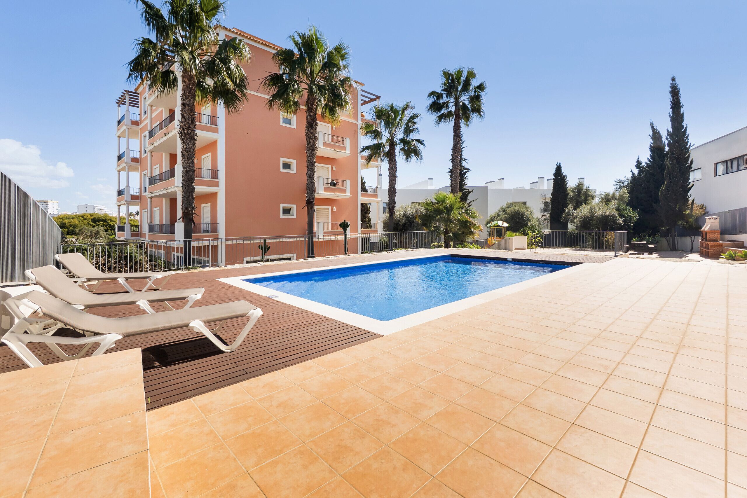 A swimming pool with lounge chairs, palm trees, and a multi-story building in the background.