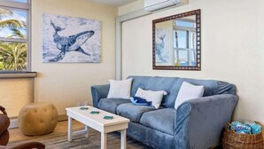 Ein Zimmer mit grauem Sofa, weißem Tisch, blauem Kunstwerk, Spiegel und einem Korbwäsche.