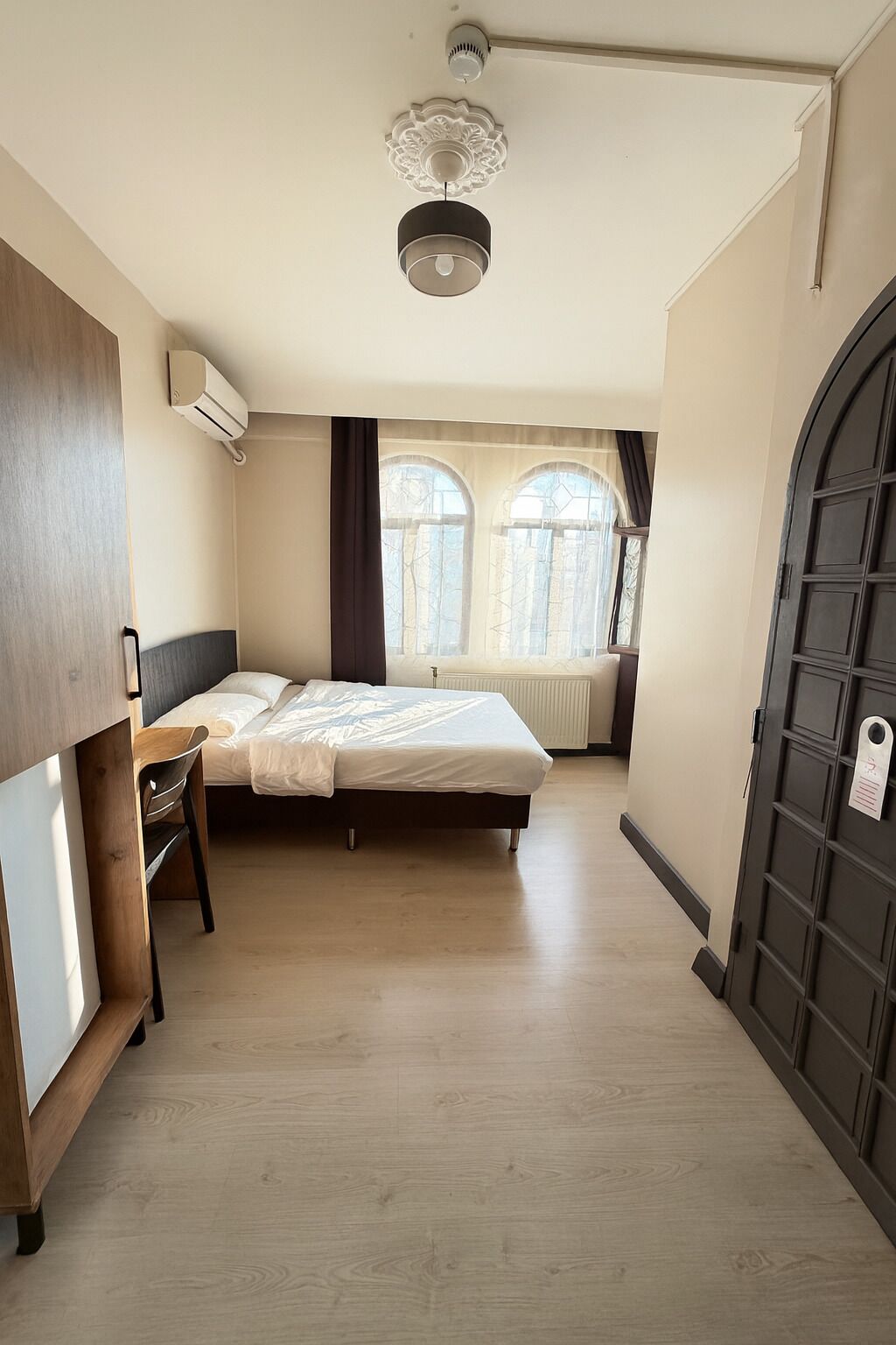 A bedroom with a bed, a desk, and a wardrobe. There is a window with curtains and a radiator.
