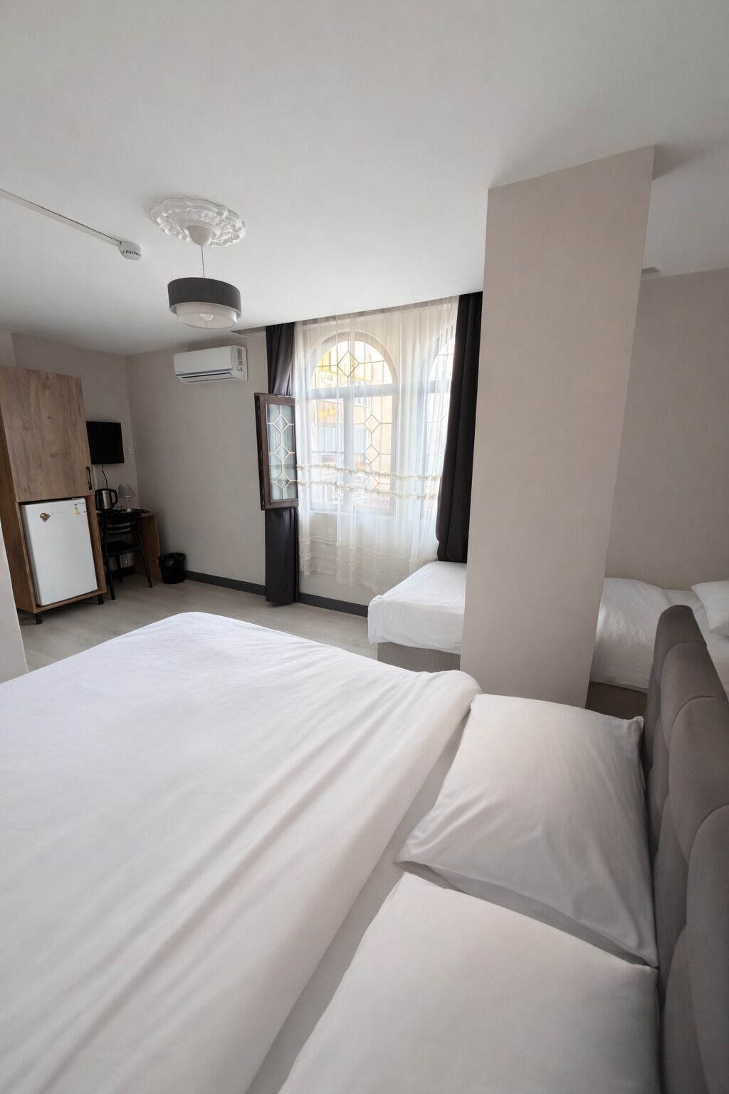 A hotel room with a large bed, a smaller bed, a desk with a chair, a wardrobe, and a window with curtains.