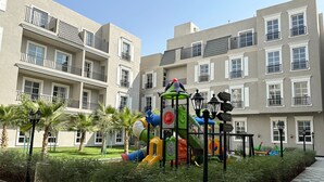 A playground with colorful equipment, surrounded by a well-maintained garden and a building with balconies. - Hittin B-3 Latest Development  AX58 (Riyadh)