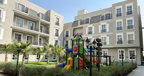 Un terrain de jeux doté d’équipements colorés, entouré d’un jardin bien entretenu et bordé d’un bâtiment avec des balcons.