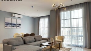 Una sala de estar moderna con un sofá modular, una mesa de centro y un comedor con sillas. Hay un ventanal con cortinas, un equipo de aire acondicionado en la pared y una obra de arte sobre el sofá.