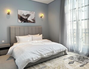 A modern bedroom with a large bed, two wall-mounted lights, a painting, and a large window with sheer curtains. - Hittin B-12 Latest Development GX58 (Riyadh)