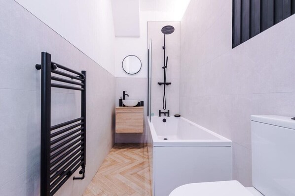 A modern bathroom with a black towel rack, a white bathtub, a wooden vanity with a sink, and a toilet. - Modern & Chic Four Bedroom Home (Manchester)