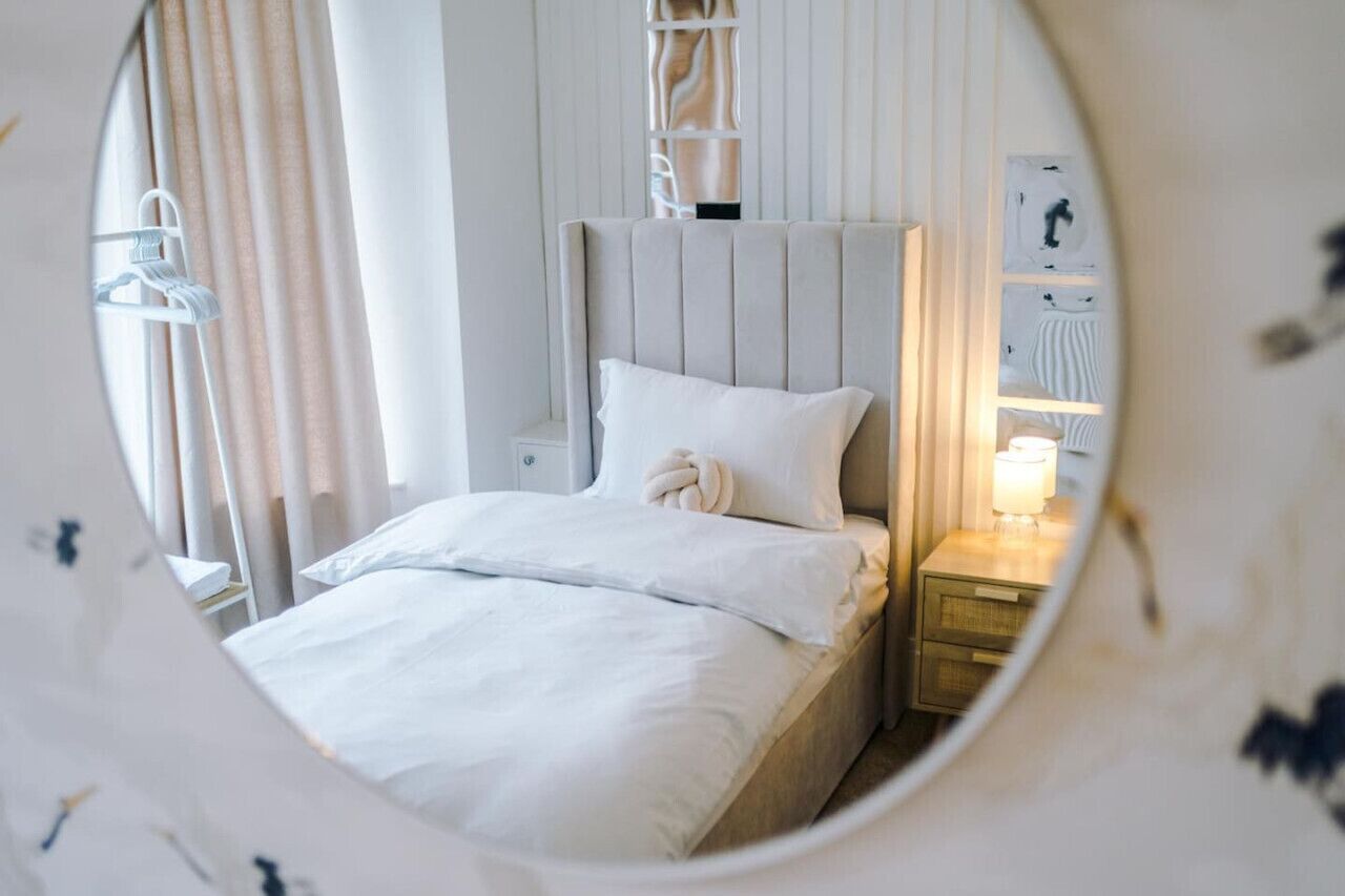 A neatly made bed with white linens and a headboard, a bedside table with a lamp, and a wall-mounted shelf with decorative items.
