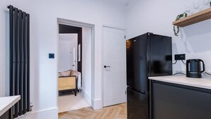 Cocina compacta con refrigerador negro, cafetera en la encimera y puerta que da a un espacio de estar con una silla de mimbre.