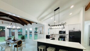 A modern kitchen with a large island, white cabinetry, and a black countertop. There is a dining area with a wooden table and chairs. - Private 30A Retreat-Large Yard-Hot Tub-2 KING Suites-Fire Pit-Boat & RV parking. (Santa Rosa Beach)