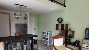 A modern kitchen with a black countertop, stainless steel refrigerator, and a dining area with stools. There is a wooden chair with a cushion and a small cabinet with shelves. - Les Myrtes Studio Style Atelier (Cuers)