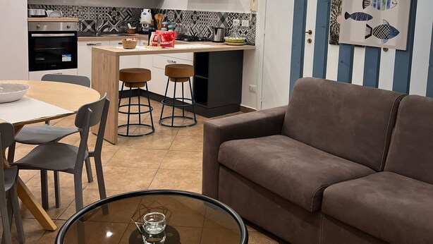 A modern kitchen with a dining area, featuring a sofa, a glass table, and a kitchen island with stools.