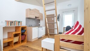 A compact living space with a kitchenette, a wooden ladder, a white sofa with a red and white striped pillow, and a wooden shelf unit. - Alpen-hostel - Apartment Mittagsonne - Buxacher Straße 23 mit Motoradvermietung (Memmingen)