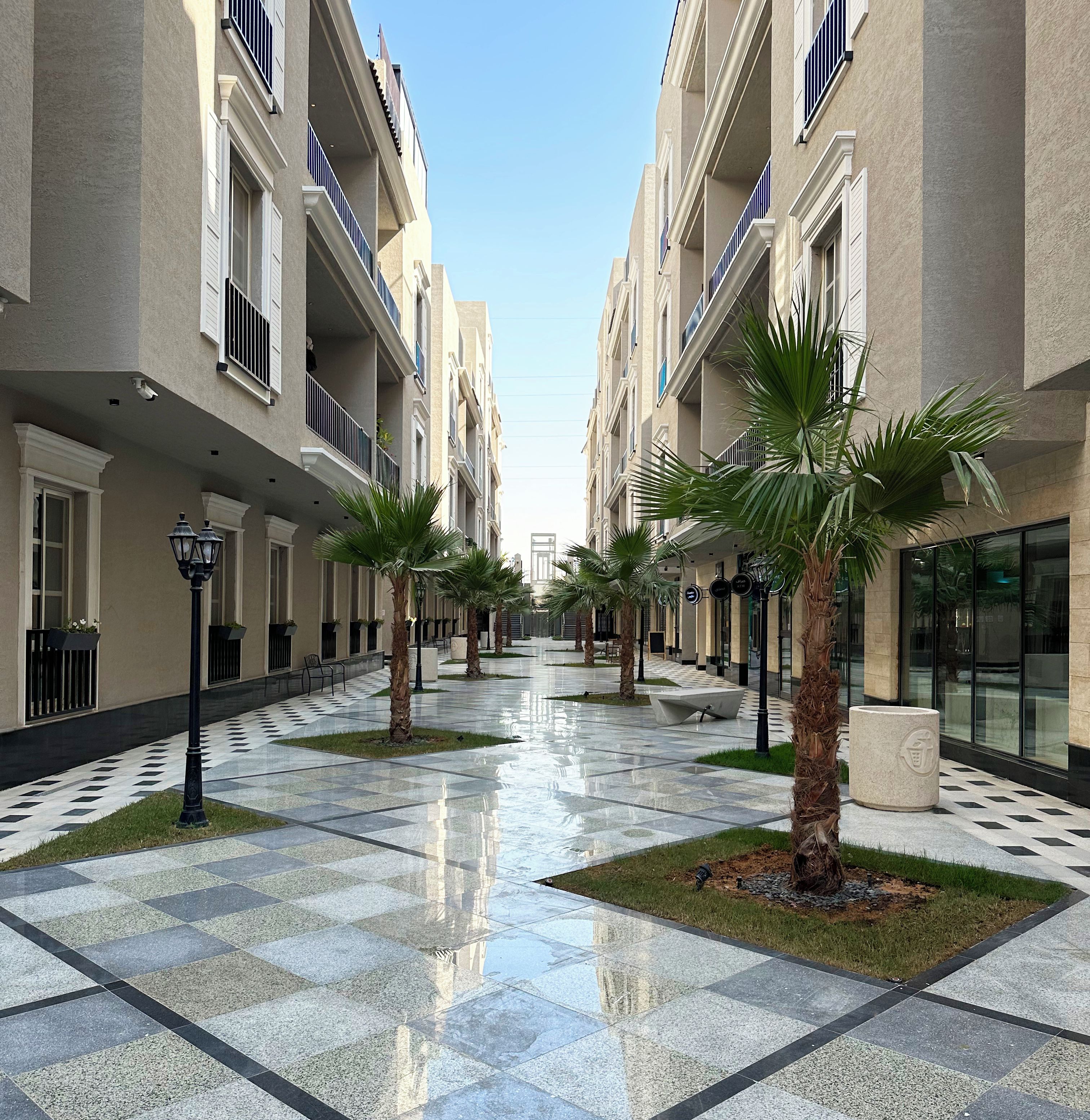 A corridor with a checkered floor, palm trees, and buildings with balconies.