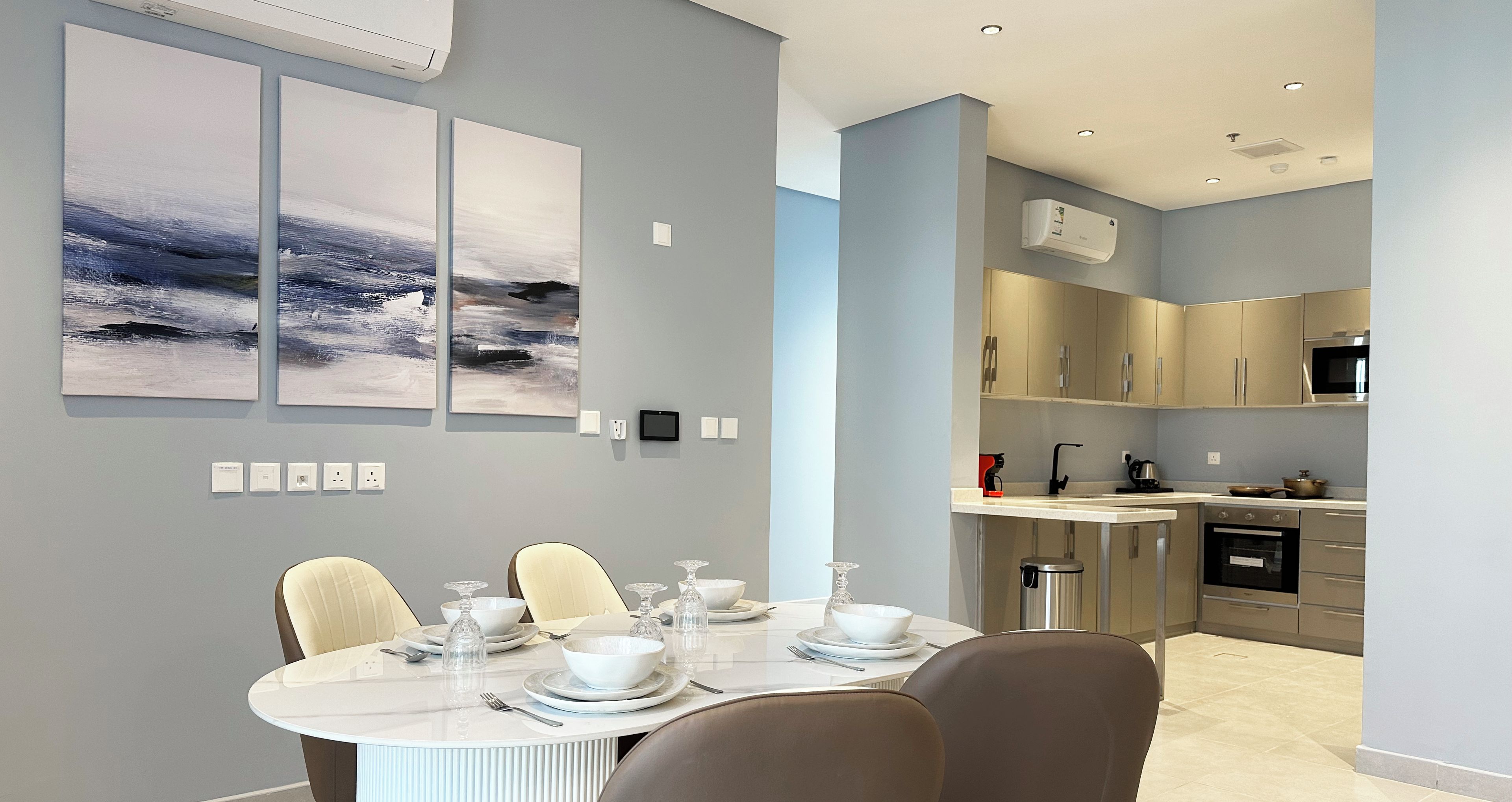 A modern dining area with a round table set for four, a kitchen with stainless steel appliances, and a grey wall with three abstract paintings.