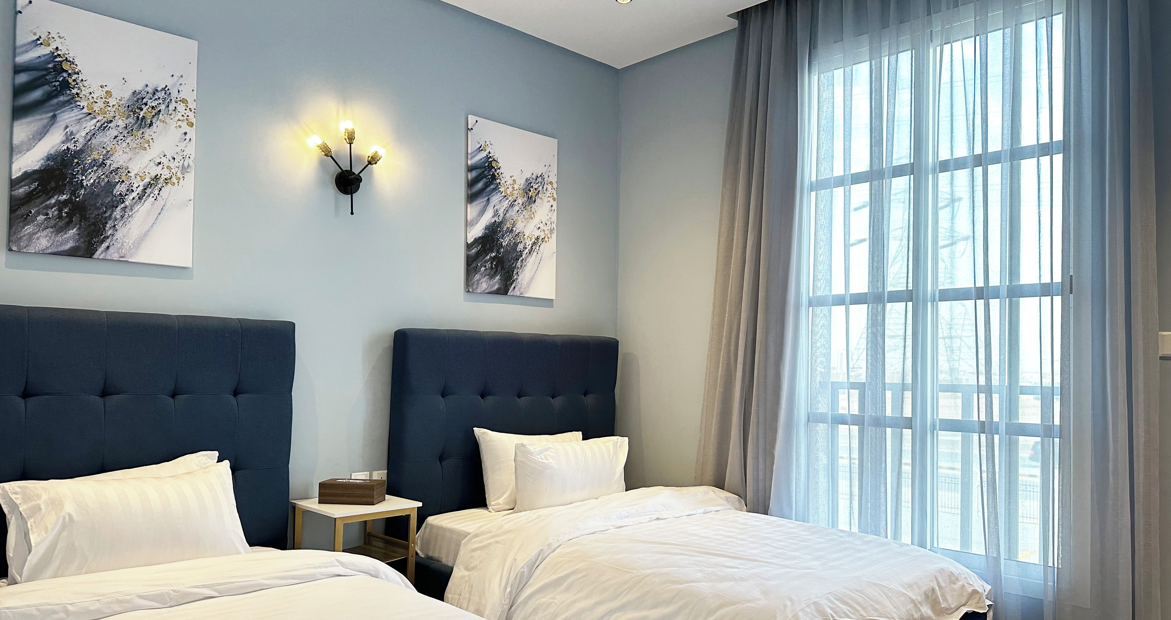 A hotel room with two beds, a nightstand, a wall-mounted light, and large windows with sheer curtains.