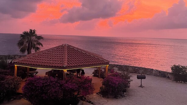 Un belvédère doté d’un toit en tuiles, entouré de fleurs violettes, offrant une vue sur une plage au coucher du soleil.