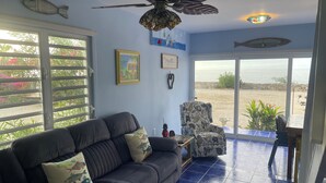 Un salon avec un canapé foncé, un fauteuil à motifs, un ventilateur de plafond et une vue sur la mer par de grandes fenêtres.
