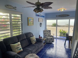 A living room with a dark sofa, a patterned armchair, a ceiling fan, and a view of the sea through large windows.