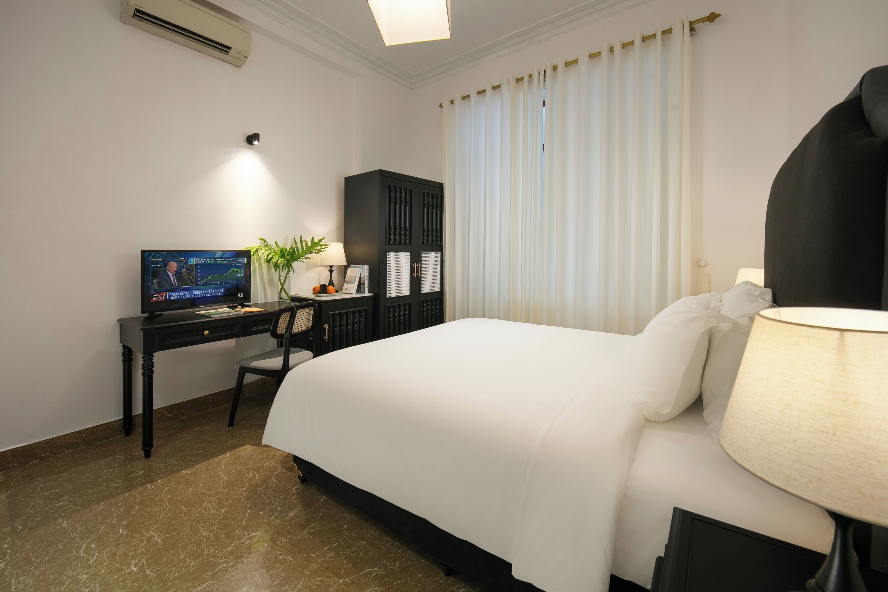 A hotel room with a large bed, a desk with a computer, a chair, a lamp, and a window with curtains.