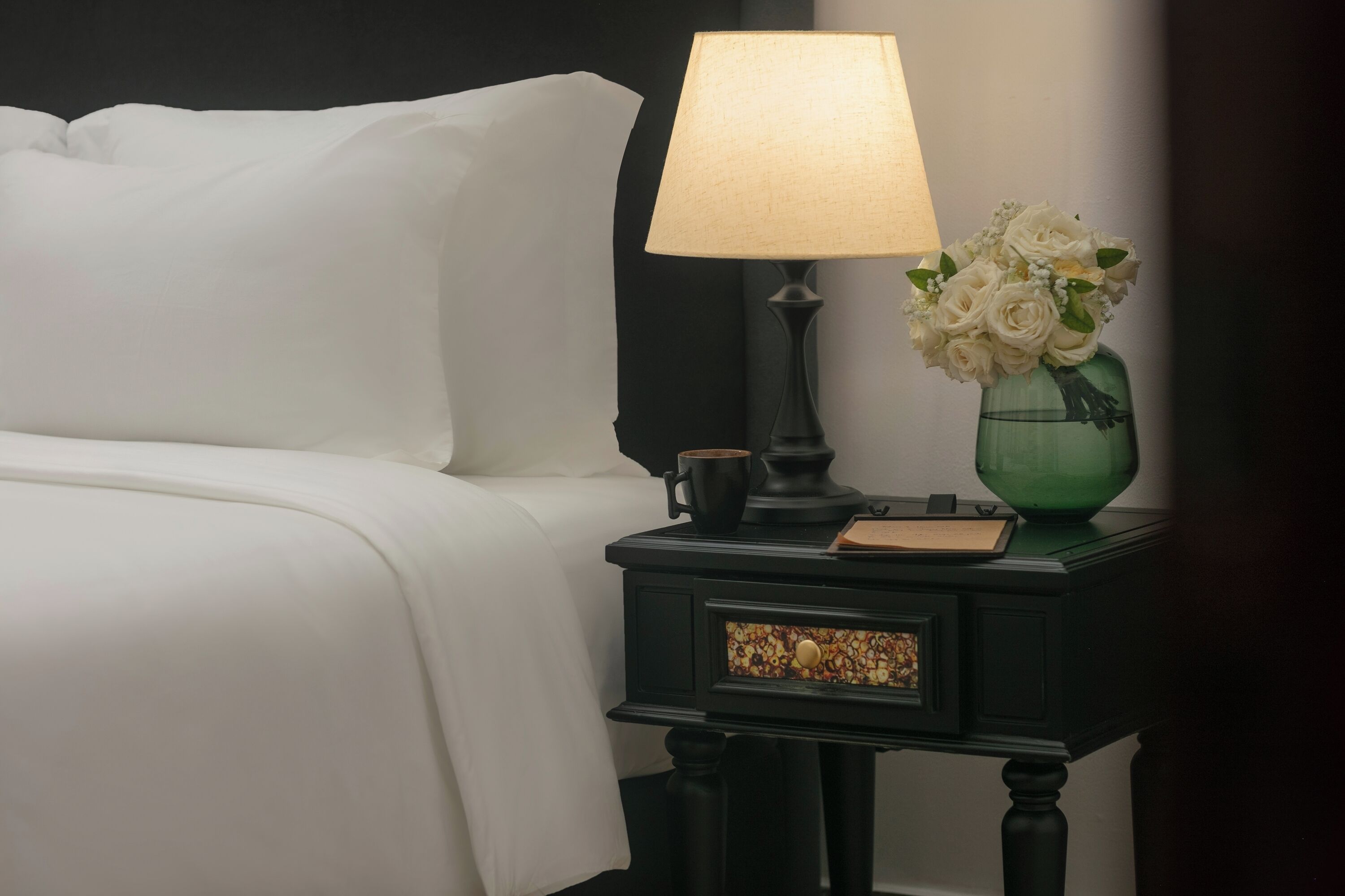 A neatly made bed with white linens, a black nightstand with a lamp, a green vase with white flowers, and a small book.