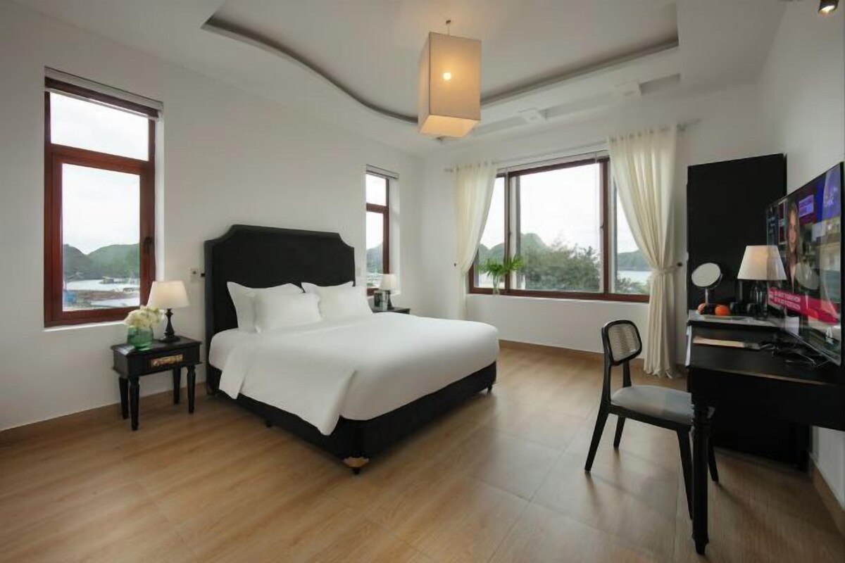 Une chambre d’hôtel équipée d’un lit, d’un bureau, d’une chaise, d’une télévision et de grandes fenêtres donnant sur un paysage agréable.