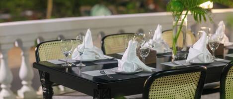 Una mesa de comedor dispuesta con servilletas blancas, copas de vino y platos, con sillas y una planta en maceta.