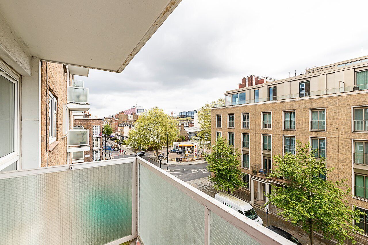 A residential building with multiple windows, a balcony, and a street view with trees and other buildings.