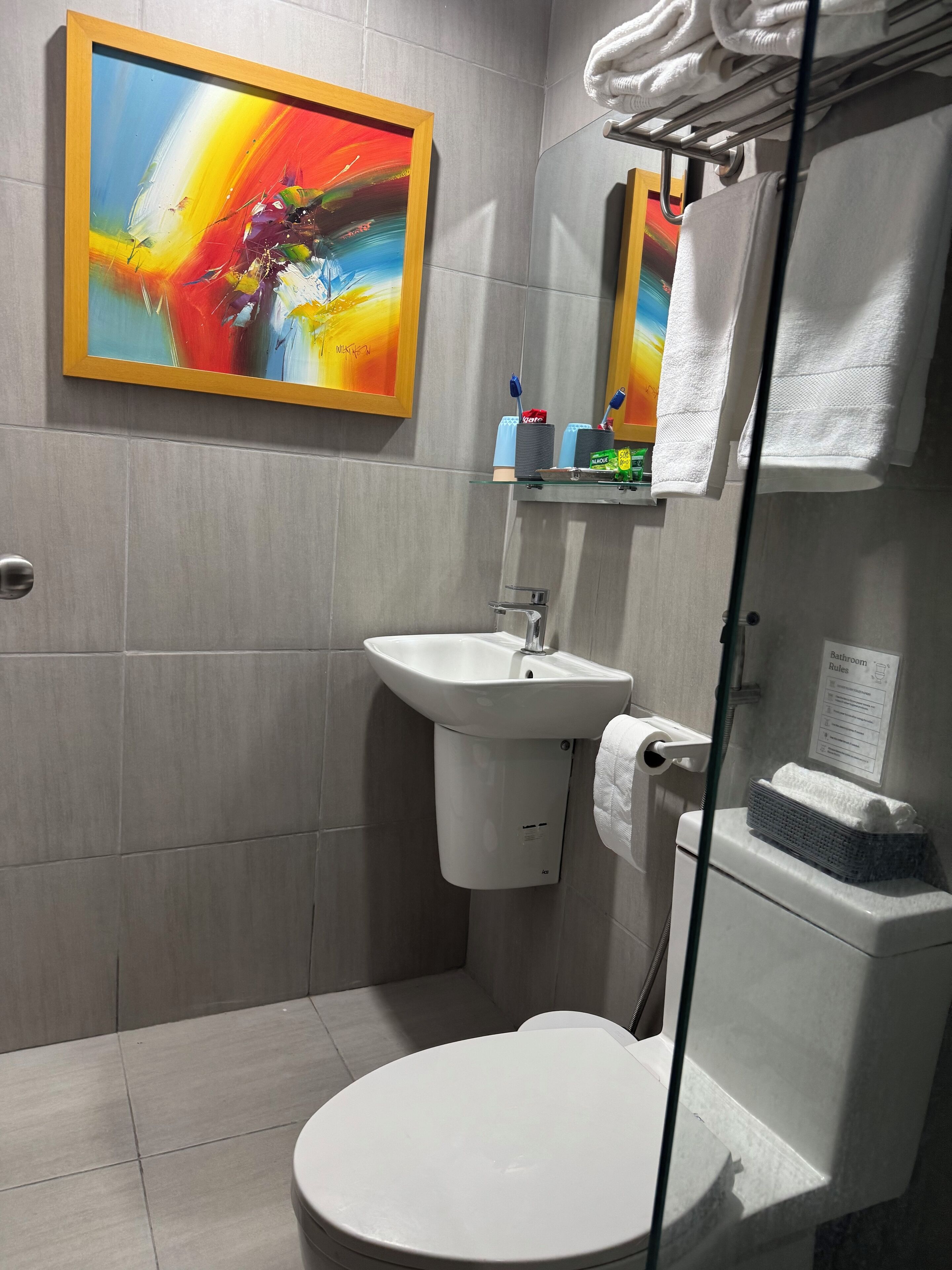 Une salle de bain moderne avec un lavabo blanc, des toilettes et une étagère en verre au-dessus du lavabo où sont rangés les articles de toilette.