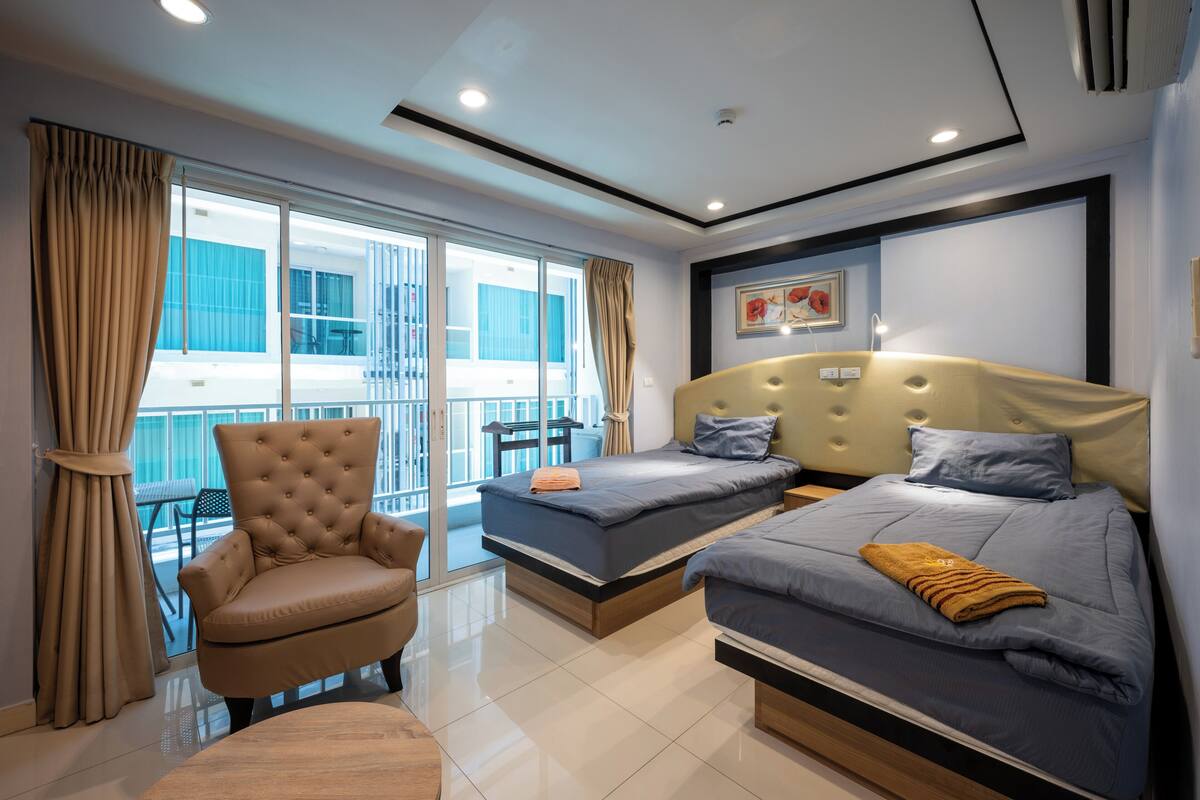 Una habitación de hotel moderna con un amplio ventanal corredero a un balcón, un sillón capitonado, dos camas individuales y una mesa de centro de madera.