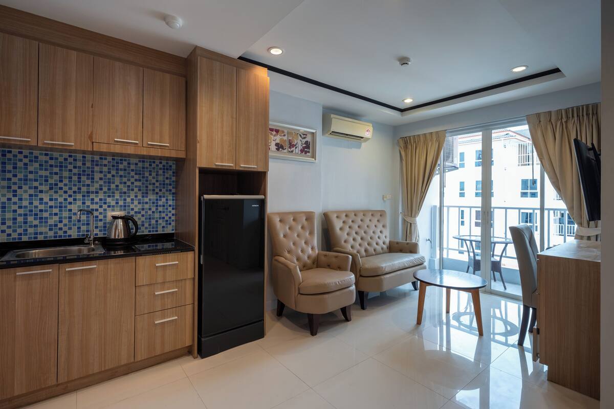 Una cocina moderna con armarios de madera, un refrigerador negro y un salpicadero de azulejos. Hay dos sillones color beige y una pequeña mesa en una zona de estar luminosa con un ventanal grande.