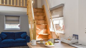 A room with a blue sofa, a wooden staircase, a dining table with a bowl of fruit, and a window with blinds. - 1 bedroom accommodation in Padstow (Padstow)