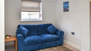 A room with a blue sofa, a small table with a bowl of fruit, a window with blinds, a painting on the wall, and a power outlet. - 1 bedroom accommodation in Padstow (Padstow)