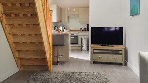 A modern kitchen with a built-in oven, a dining area with a bar stool, a flat-screen TV on a wooden stand, and a staircase leading to an upper level. - 1 bedroom accommodation in Padstow (Padstow)