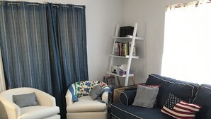 Ein Wohnzimmer mit weißem Sessel, einer Couch mit Kissen mit amerikanischer Flagge, einem weißen Couchtisch mit Fernbedienungen, einem Bücherregal und einem Fenster mit Vorhängen.