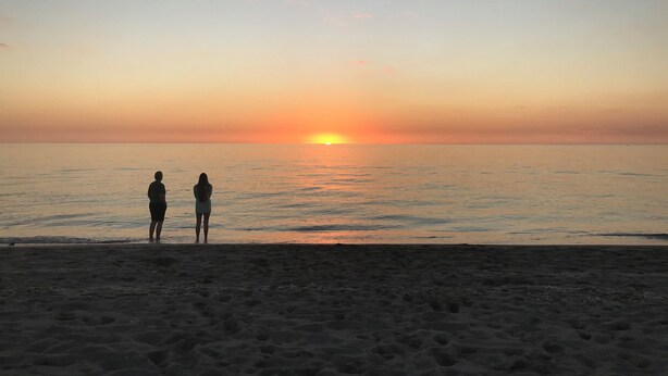 Zwei Personen stehen am Strand und schauen dem Sonnenuntergang entgegen.