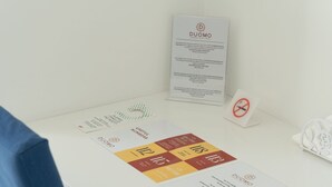 Un escritorio blanco con una silla azul, un letrero con la inscripción 'DUOMO', un cartel de 'no fumar' y una tarjeta con los textos 'NUMEROS ÚTILES' y 'DUOMO'.