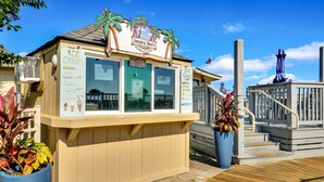 A small ice cream stand with a menu board, located on a wooden deck with a railing and potted plants.