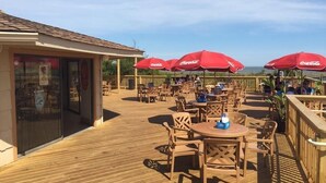 An outdoor patio with wooden tables and chairs, red umbrellas, and a view of the ocean.