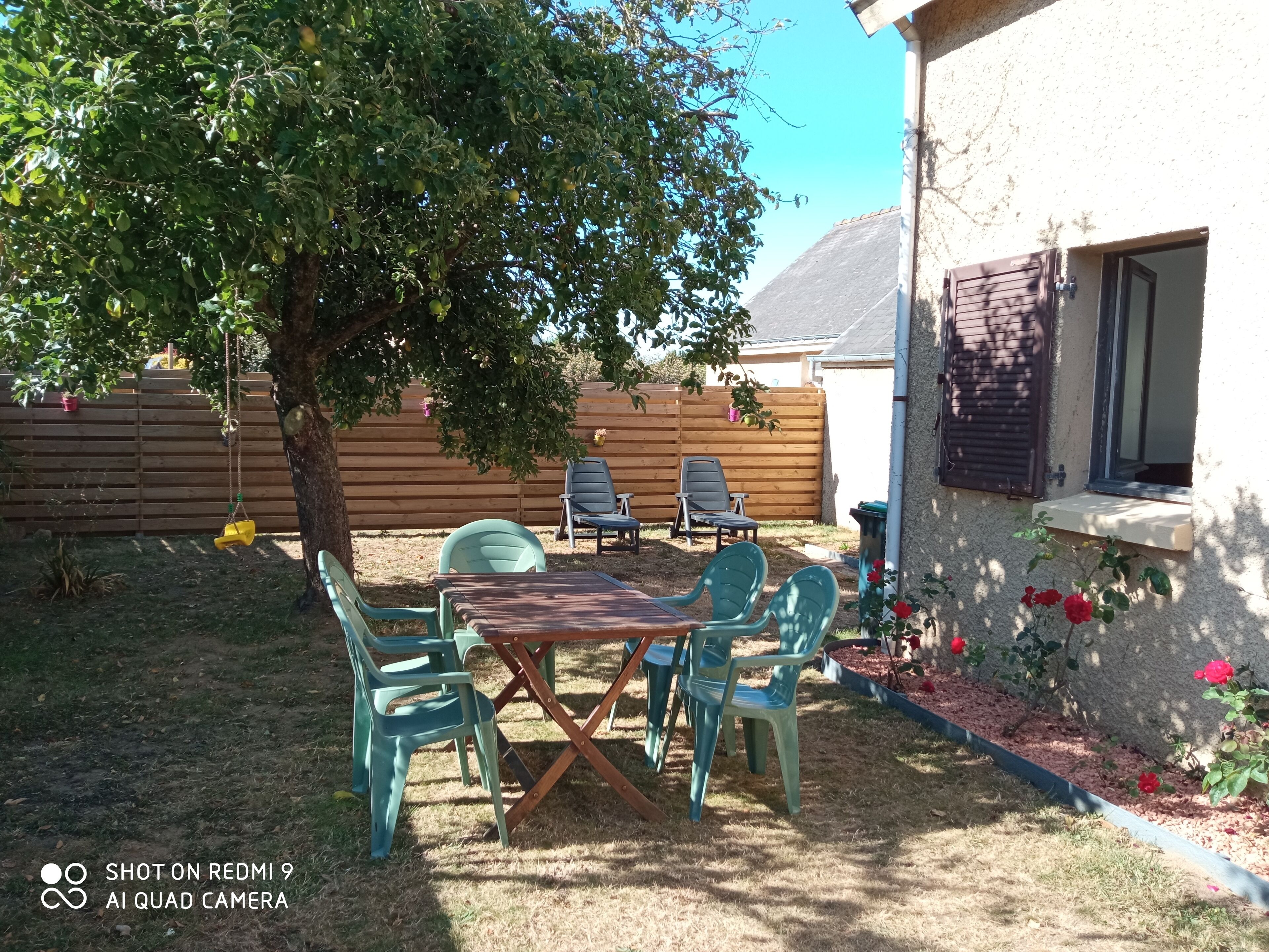A backyard with a wooden table and four chairs, a swing, a wooden fence, and a potted plant with red flowers.