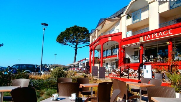An outdoor cafe area with tables and chairs, a red building with 'LA PLAGE' signage, and a clear blue sky.