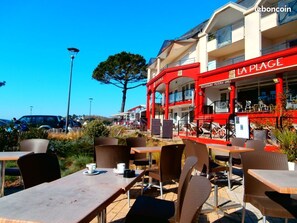 Un área de café al aire libre con mesas y sillas, un edificio rojo con el letrero 'LA PLAGE' y un cielo azul despejado.