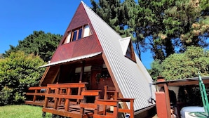 A small, triangular wooden cabin with a sloped roof, a porch with wooden benches, and a car parked nearby. - CasaAlpina (Tandil)