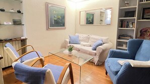Un salon avec un canapé blanc, des fauteuils bleus, une table basse en verre et une bibliothèque.