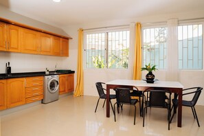 A kitchen with wooden cabinets, a washing machine, a dining table with chairs, and large windows with curtains.