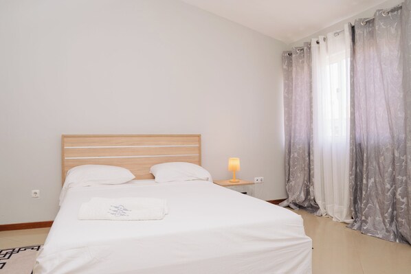A neatly made bed with white linens and a pillow with a floral design, a bedside table with a lamp, and a window with grey curtains.