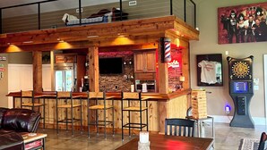 Un salón espacioso con ventilador de techo alto, una barra de madera con taburetes, una zona de cocina y un área de estar con mesa y sillas.