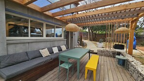 A covered outdoor seating area with a wooden deck, a gray sofa with cushions, a green table, and a yellow bench.