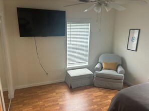 A bedroom with a ceiling fan, a TV mounted on the wall, a window with blinds, a checkered armchair, and a bedside table. - The Goodlife cottage#2 quiet and relaxing (Palm city)